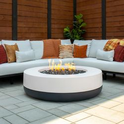 A Round Firepit with a dark bottom, and chalk colored top. It is filled with black fire glass which has flame coming out of the center. the fire pit rests between a sectional outdoor couch, nestled into a wooden fenced in patio.