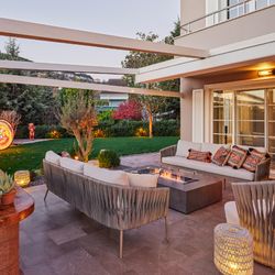 Woven outdoor lounge chairs in tan with metal legs, arranged around a rectangular fire pit table on a modern patio with pergola overhead