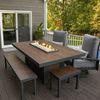 Outdoor fire pit dining table with brown wooden or material composite top, black metal frame, and built-in linear gas fire feature, paired with matching bench and c